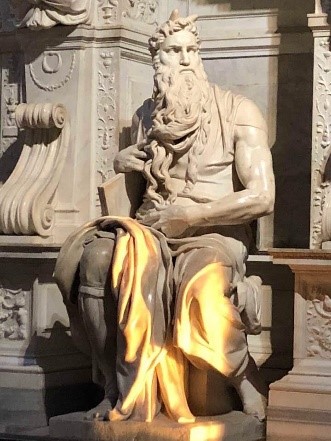 Moses by Michelangelo. Marble statue of a seated man with long beard and serious look, a tablet in one arm. Behind a wall with many details in high relief.