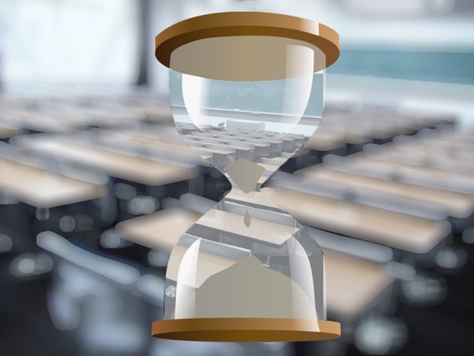 An hourglass with falling sand (most of the sand is at the bottom). In the background is a blurred photo of a classroom. You can see the room clearly through the glass of the hourglass.