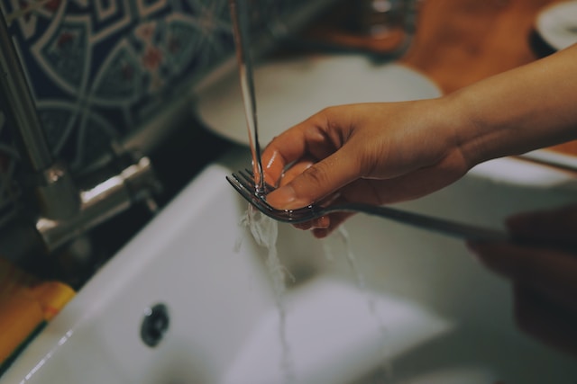 Photograph of a person washing dishes, specifically focusing on a fork. In the image, only a part of their body is visible: the hand.