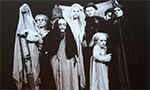 Black and white photograph of the show "Métamorphoses" by Ilka Schönbein, in which slightly macabre-looking puppets and marionettes are on a wooden platform, being manipulated by the artist.