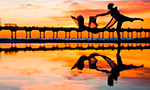 Photograph of a person dancing with a scarf in their hands, against the light of the sunset, in front of a bridge. You can see the person's reflection in the water.