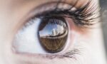 Close-up of a brown eye. In the reflection you can see a balcony screen in the foreground and, in the distance, other buildings, suggesting that the model is on the balcony of an apartment.