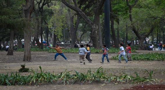 Foto de seis niños jugando con una pelota en una plaza. Es un espacio con muchos árboles y plantas.