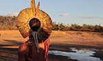 Fotografía de un hombre indígena de espaldas, mirando el horizonte. Lleva un tocado de plumas y varios adornos en el cuerpo. Está junto a un pequeño lago en un campo abierto con vegetación variada. El cielo está despejado y azul.