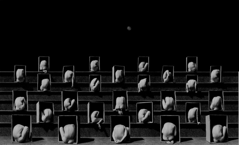 Fotografía en blanco y negro de una grada compuesta por cuerpos desnudos encogidos dentro de cajas con la luna al fondo. Fotografía en blanco y negro de una grada compuesta por cuerpos desnudos encogidos dentro de cajas con la luna al fondo.