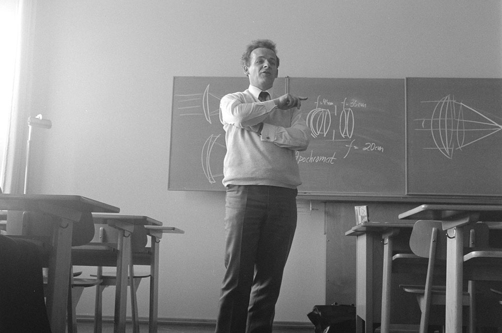 Foto em preto e branco. Professor em pé em uma sala de aula, atrás um quadro com representações óticas e ao lado cadeiras de alunos.