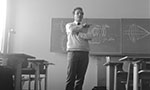 Foto em preto e branco. Professor em pé em uma sala de aula, atrás um quadro com representações óticas e ao lado cadeiras de alunos.