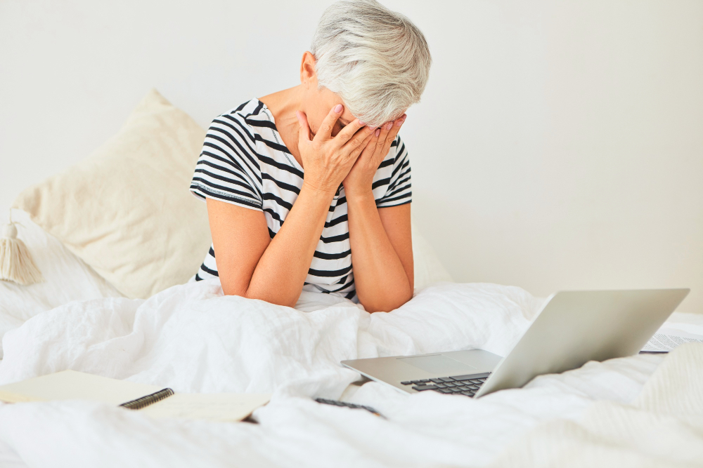 Uma mulher de meia idade, pele branca, cabelo curto e branco acinzentado veste uma camiseta listrada preta e branca. Ela está sentada de pernas cruzadas em uma cama e está de frente para um laptop. Ela está com as mãos no rosto parecendo frustrada.
