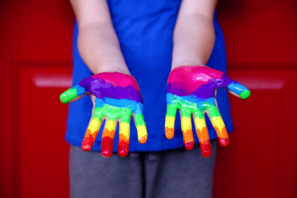Uma criança veste uma blusa azul e estende as mãos viradas pra cima, elas estão pintadas formando um arco-íris.