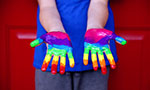 Uma criança veste uma blusa azul e estende as mãos viradas pra cima, elas estão pintadas formando um arco-íris.