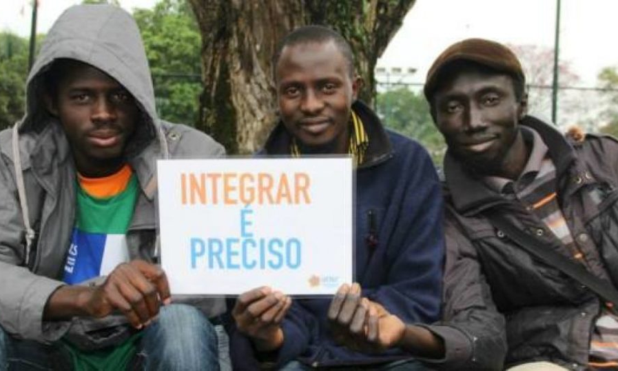 Três homens negros sentados segurando uma folha de papel com a frase "Integrar é preciso"