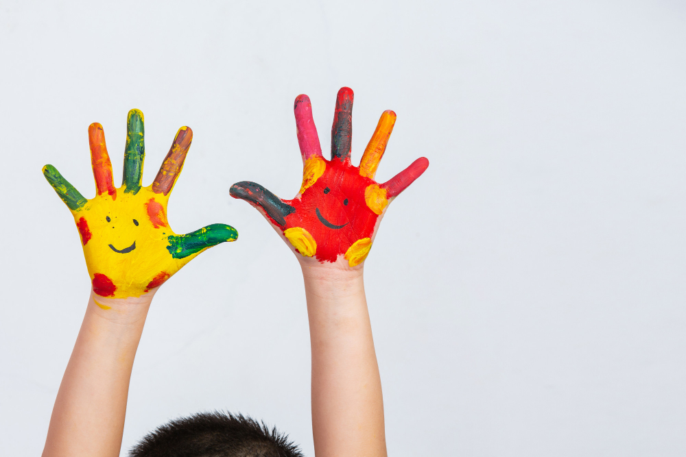 Photo. Light gray background, two forearms of a child with hands wide open and palms facing forward. Their skin is white and their hands are painted with colored paint (yellow, red, orange, pink, green, brown), each with two eyes and a smile drawn with black paint. You can also see the top of the child's head, they have short black hair.