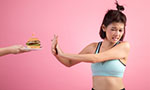 Foto retangular horizontal. Uma mulher adulta recusa um hambúrguer. Ela faz uma careta, estende os braços na direção do hambúrguer e sobrepõe as duas mãos. Ela usa roupa de ginástica. Fundo rosa difuso.