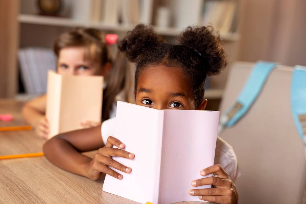 Duas crianças sentadas em frente a uma mesa com livros cobrindo parte do nariz e da boca