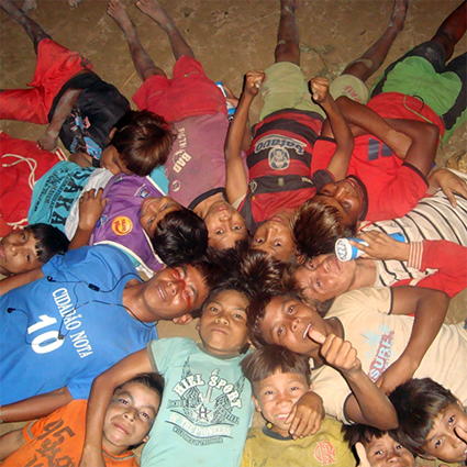 Foto de meninos indígenas deitados no chão. Eles foram um círculo com as cabeças próximas no centro. Eles parecem estar se divertindo.