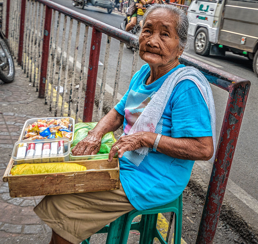 Foto: senhora idosa sentada em um banquinho de plástico na rua com uma caixa no colo. Na caixa: cigarros e salgadinhos que ela parece estar vendendo. A expressão dela é neutra, mas também demonstra cansaço.