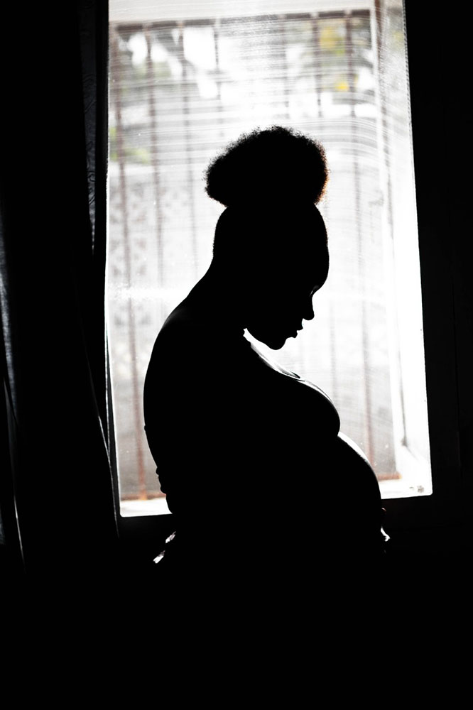 Foto em alto contraste. A sombra de uma mulher negra grávida posando de perfil. Ela olha para baixo. Atrás, uma janela com muita iluminação e o ambiente todo escuro.