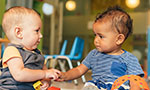 Foto: dois bebês olhando um para o outro e segurando as mãos. O bebê da esquerda segura um bloco de brincar.