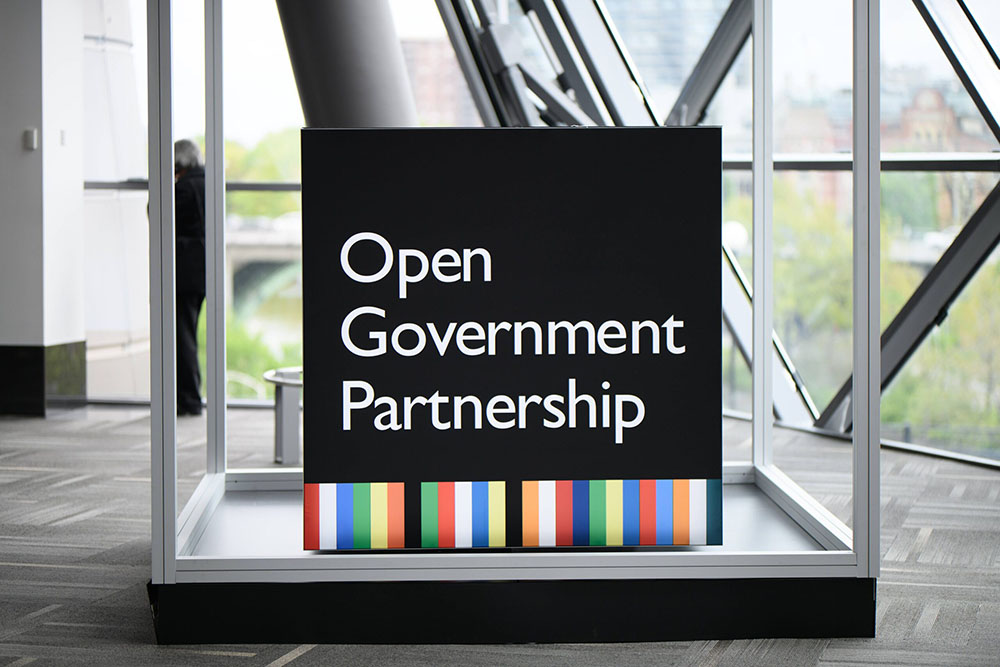 Foto de um quadro preto com o texto "Open Government Partnership". Uma faixa listrada e colorida na parte de baixo. O quadr está dentro de um aquário vazio e retangular. No fundo, uma sala ampla e clara com paredes envidraçadas.