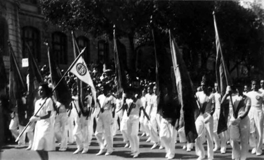Foto em preto e branco. Um grupo de pessoas desfilando na rua. Elas usam roupas brancas e levam bandeiras. No fundo, um prédio comprido e árvores.