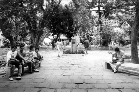 Foto em preto e branco. Uma passagem em uma praça. Na esquerda, um grupo de homens sentado em um banco. No centro, mulher andando ao lado de uma estátua que está no meio da praça. Na direita, um homem sentado lendo um livro. No fundo, muitas árvores e grama.