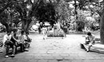 Foto em preto e branco. Uma passagem em uma praça. Na esquerda, um grupo de homens sentado em um banco. No centro, mulher andando ao lado de uma estátua que está no meio da praça. Na direita, um homem sentado lendo um livro. No fundo, muitas árvores e grama.
