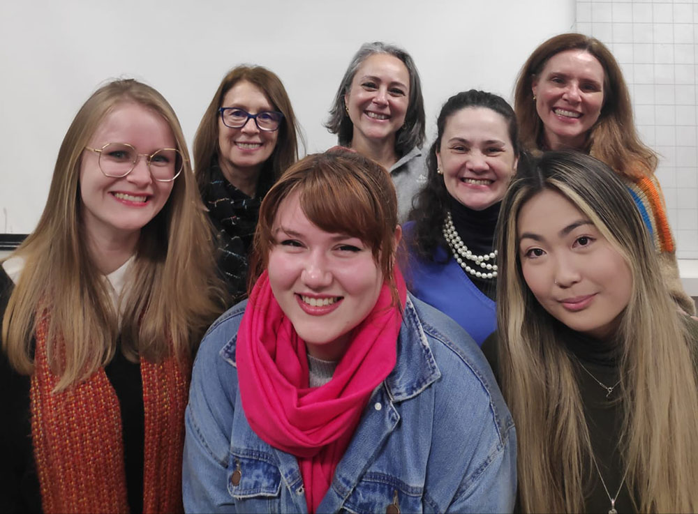 Foto: sete mulheres de idades variadas estão próximas umas das outras, sorrindo e olhando para a câmera. Parece ter uma lousa no fundo da sala.