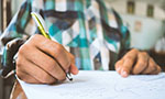 Foto: mão segurando caneta e fazendo anotações em um papel. É uma pessoa adulta e o ambiente parece ser uma sala de aula.