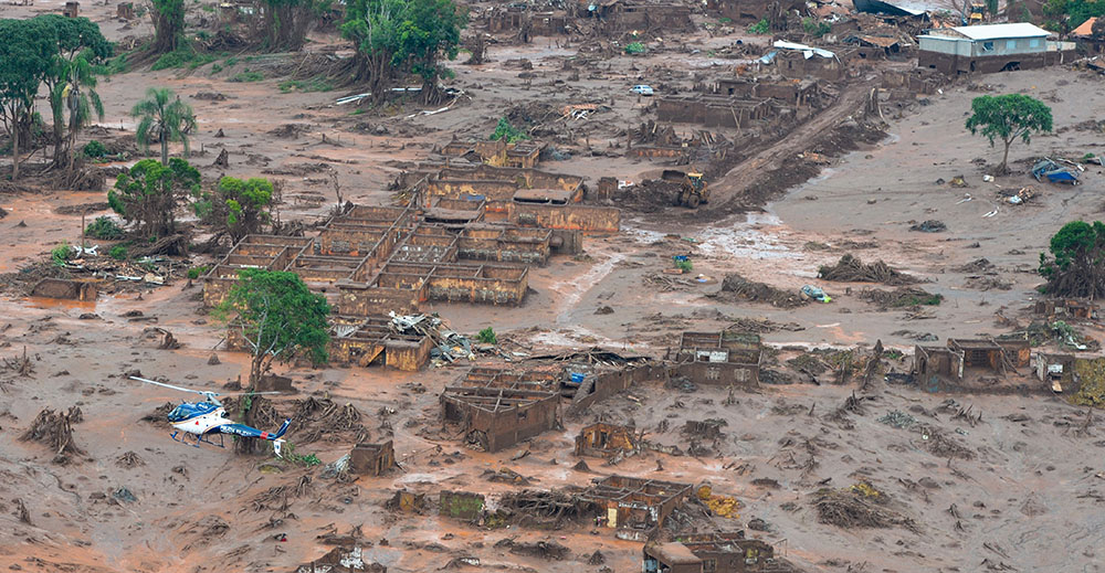 Foto aérea tirada em Mariana, Minas Gerais (2015) após o rompimento de uma barragem. A foto mostra uma região plana coberta por lama, casas destruídas, árvores isoladas e um helicóptero sobrevoando.