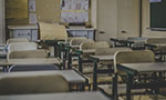 Foto de uma sala de aula com várias cadeiras enfileiradas. No fundo, armários e desenhos colados na parede. O tom da foto está meio escurecido, dando a impressão de ser um lugar abandonado.