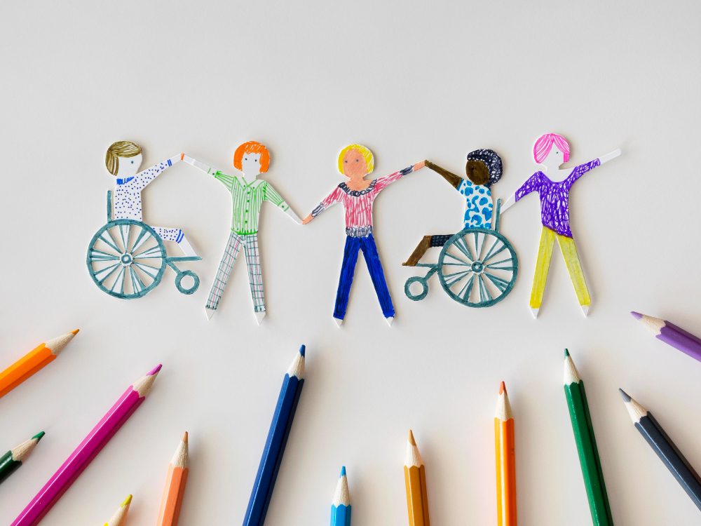 Composition. A drawing made with colored pencils has been cut out in the shape of people. They are holding their hands in a line. Two people are wheelchair users. In the lower part, colored pencils are arranged pointing at the drawing. Solid white background.