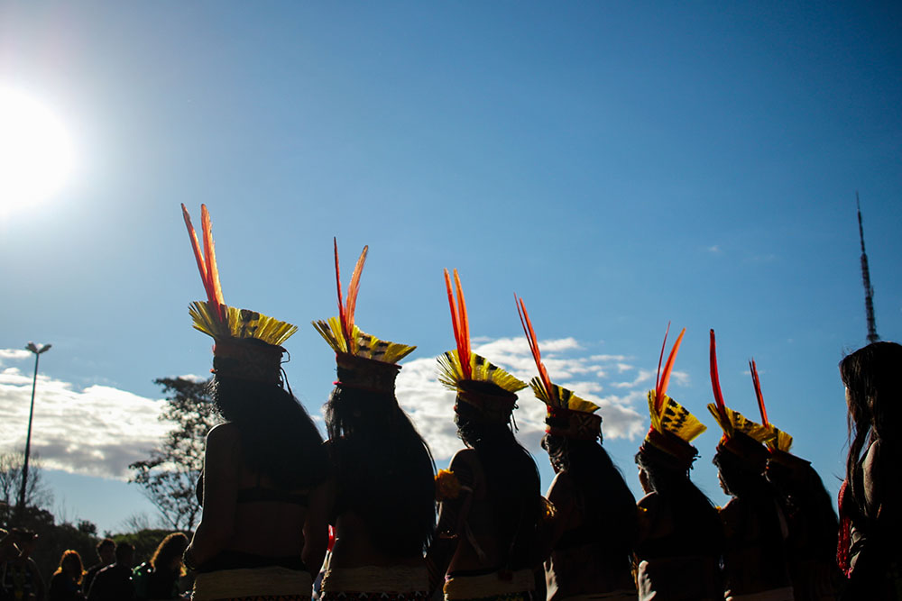 Foto da Marcha das Mulheres Indígenas, Brasília - DF (2019). Oito mulheres indígenas de costas. É possível ver apenas a silhueta e seus cocares. No fundo, céu azul com poucas nuvens, o sol, algumas árvores e um grupo de pessoas.