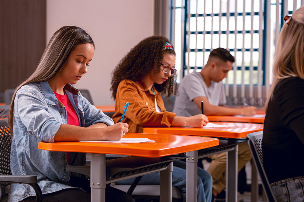 Foto de quatro pessoas em uma sala de aula. Cada um segura uma caneta e escreve em um papel. Elas parecem estar concentradas e sérias.