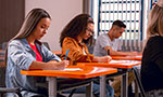Foto de quatro pessoas em uma sala de aula. Cada um segura uma caneta e escreve em um papel. Elas parecem estar concentradas e sérias.