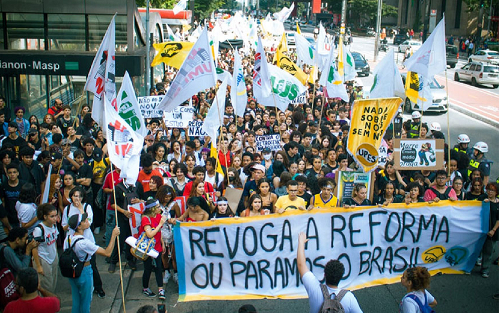Foto de um protesto. Pessoas jovens ocupam duas faixas da Av. Paulista ao lado da estação Trianon-Masp. Elas seguram bandeiras e na frente levam um cartaz dizendo "Revoga a reforma ou paramos o Brasil".
