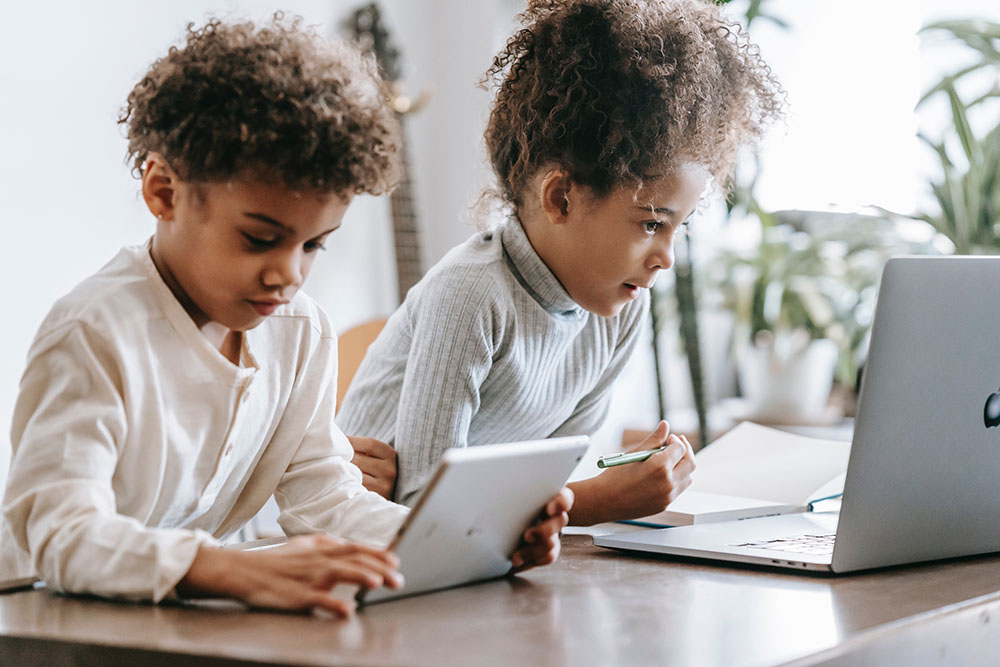 Foto de duas crianças sentadas em uma mesa. Uma delas usa um tablet e a outra olha para a tela de um notebook enquanto segura um lápis próximo de um caderno.