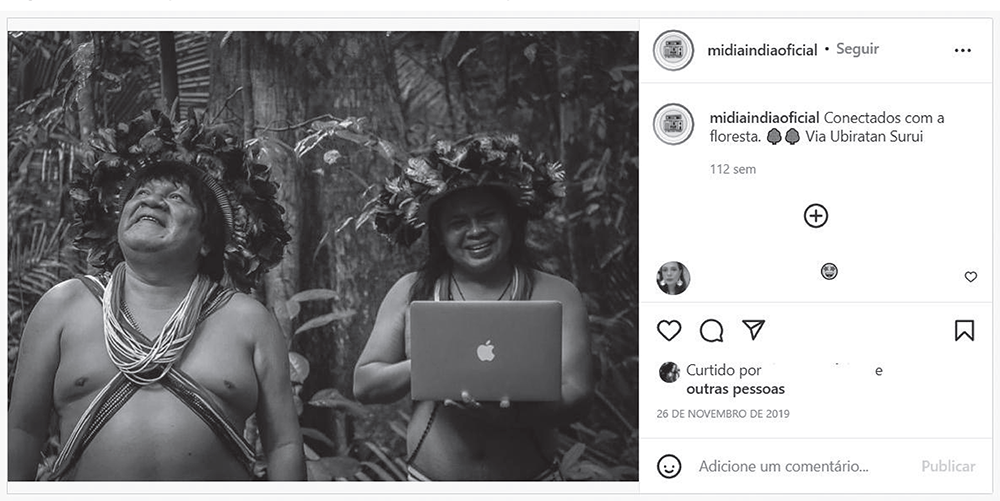 Print de uma foto no Instagram. O nome do perfil é "midiaindiaoficial". Na foto, duas pessoas indígenas estão sorrindo. no fundo, parte de uma floresta. Uma das pessoas está segurando um notebook. Na legenda, "Conectados com a floresta - via Ubiratan Surui". Publicada em 26 de novembro de 2019.