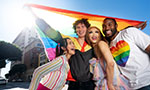 Foto quatro pessoas jovens sorrindo. Elas estão juntas posando para a foto e uma delas segura uma bandeira de arco-íris.