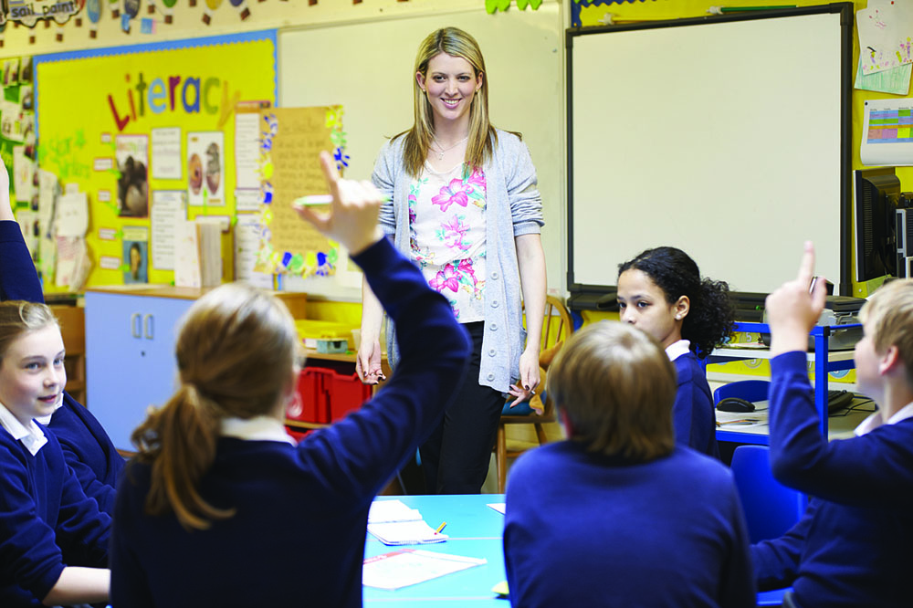 Foto de uma sala de aula. A professora está em pé sorrindo para os alunos. Dois deles estão com uma das mãos levantadas.