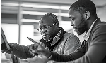 Dois homens negros sentados diante de um computador. Ambos, o mais velho e o mais novo, estão apontando para o computador. A expressão facial dos dois é de seriedade de concentração.