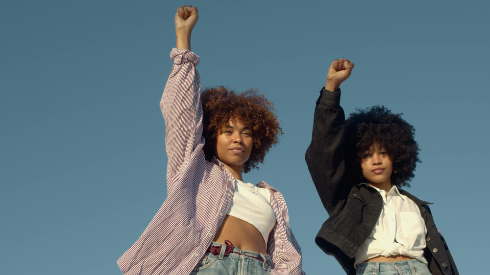 Foto de duas mulheres negras olhando para a câmera. Elas estão com o punho levantado e com expressão neutra. As duas tem cabelo afro. No fundo, um céu azul.