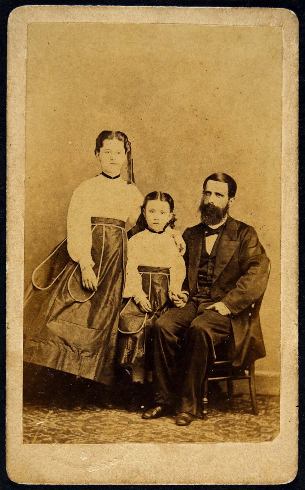 Um homem de barba está sentado, acompanhado por duas meninas. A mais nova está ao seu lado, enquanto a mais velha fica ao lado da mais nova, ambas em pé. Os três estão vestidos com trajes de época que remetem ao estilo da época retratada na imagem.