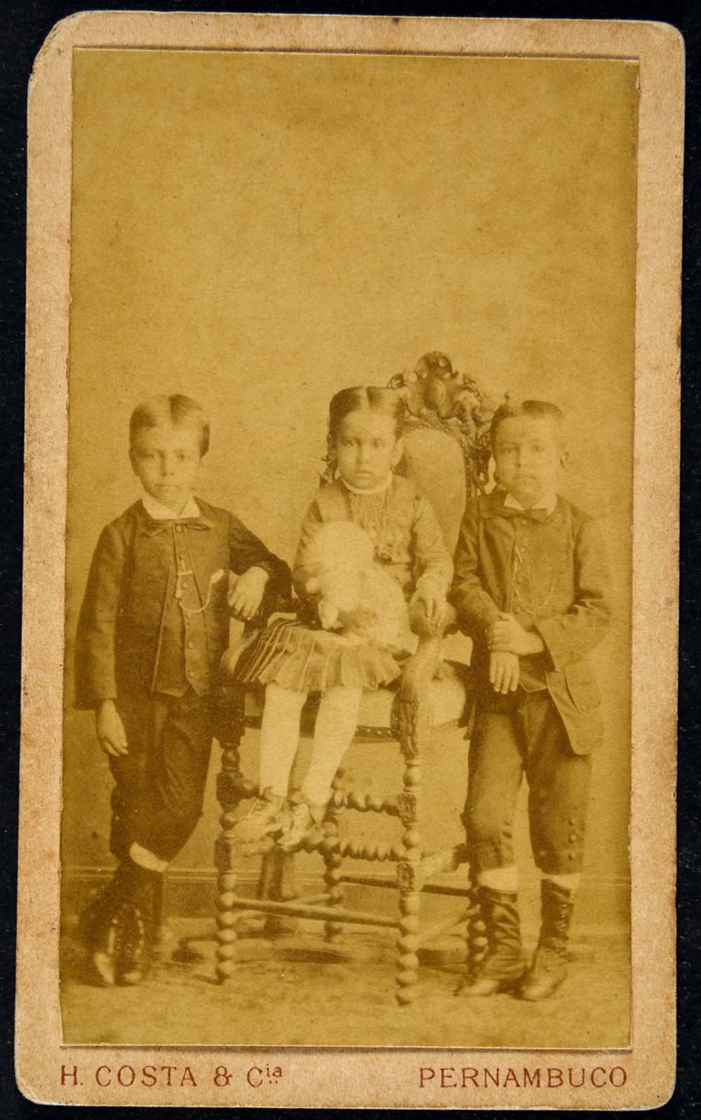 Três crianças: uma menina está no centro, sentada e segurando um brinquedo, enquanto dois meninos estão em pé, um em cada lado dela.