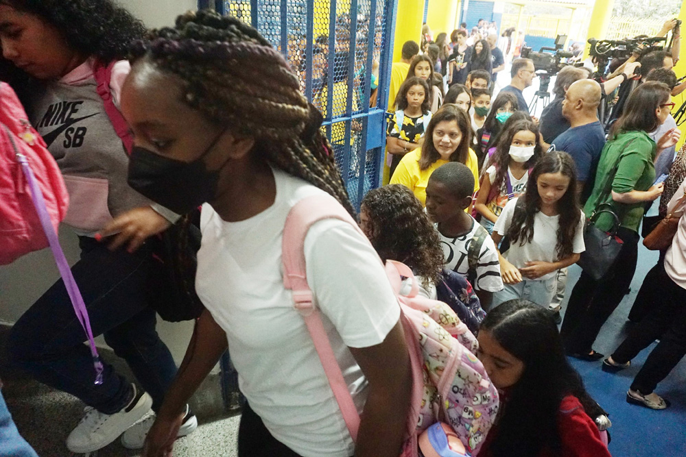 Crianças entrando na escola. A imagem destaca uma jovem estudante negra, usando uma máscara preta e uma mochila de cor rosa estampada.