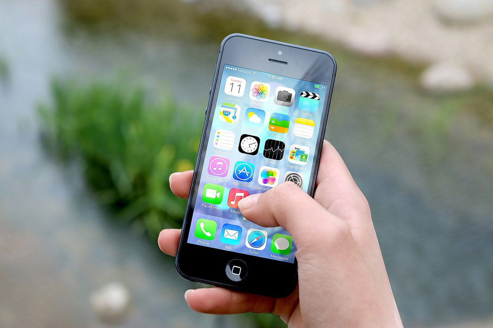 Mão segurando um celular cuja tela mostra diversos ícones de aplicativos. O fundo da imagem está desfocado.