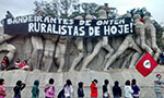 Foto do Povo Guarani em manifestação no Monumento às Bandeiras, uma estátua com vários homens enfileirados. Sobre o monumento, um cartaz dizendo “bandeirantes de ontem, ruralistas de hoje!”. Ao redor, aproximadamente 17 pessoas – uma delas leva a bandeira do MST.