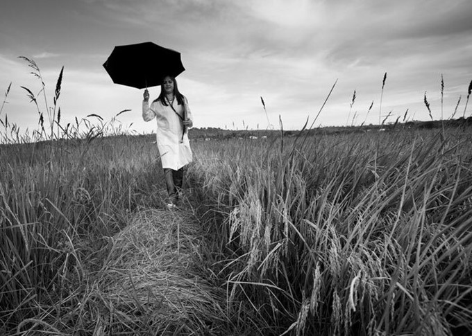 Mulher, vestida com jaleco, segurando uma prancheta e um guarda-chuva enquanto caminha sobre um campo gramado.