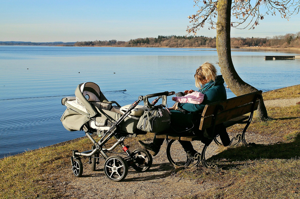 Mulher loira, vestindo roupas de frio, sentada em um banco à beira de um rio, segurando um bebê nos braços. À esquerda da mulher, encontra-se um carrinho de bebê, e à direita, uma árvore.