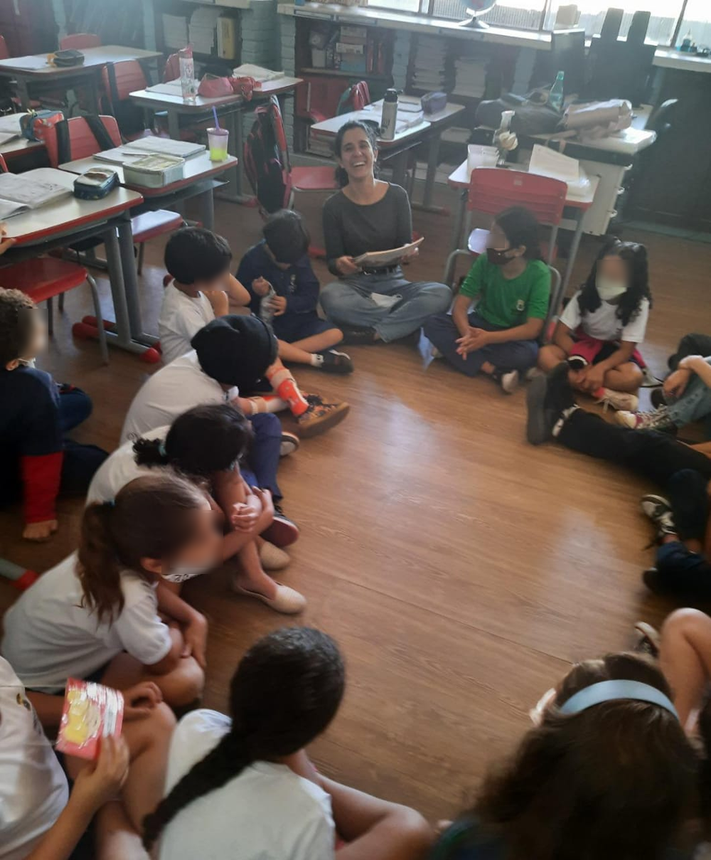 Professora e alunos sentados no chão de uma sala de aula, em círculo. A docente está sorrindo, com um livro em mãos.