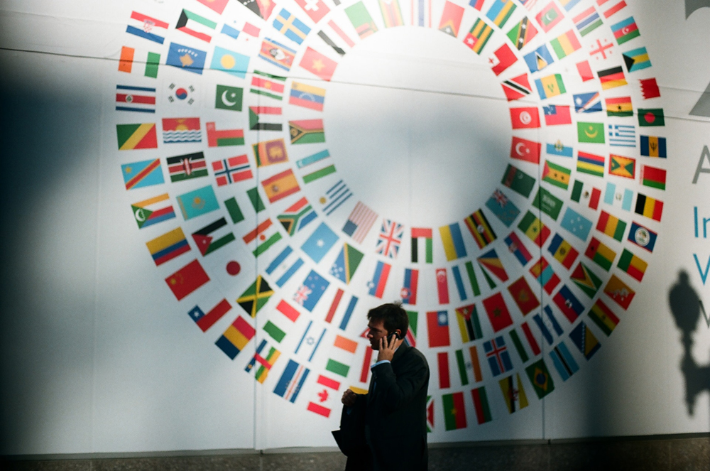 Homem vestindo terno preto, visto de perfil, falando ao celular, em frente a uma parede branca onde as bandeiras de diversos países estão dispostas em um círculo, deixando um espaço vazio no meio.